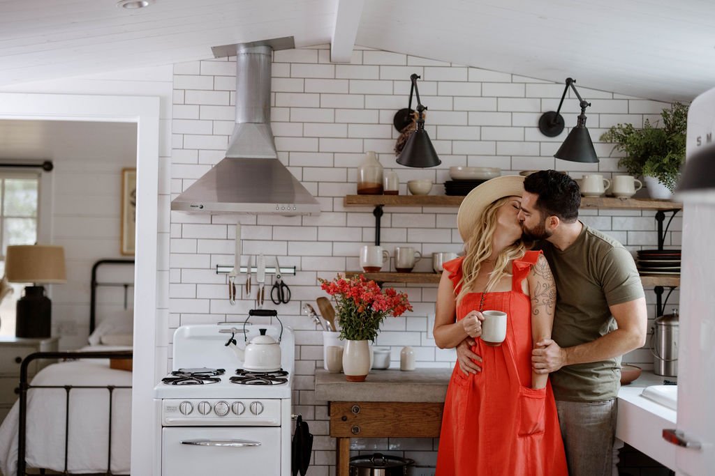 A couple sharing a kiss over morning coffee in a sunlit kitchen filled with handmade ceramics